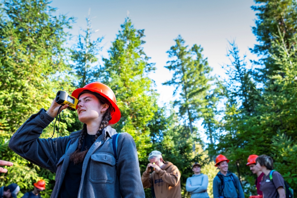 A photo of a student wearing a bright orange safety helmet, while looking through a monocular during a forestry class