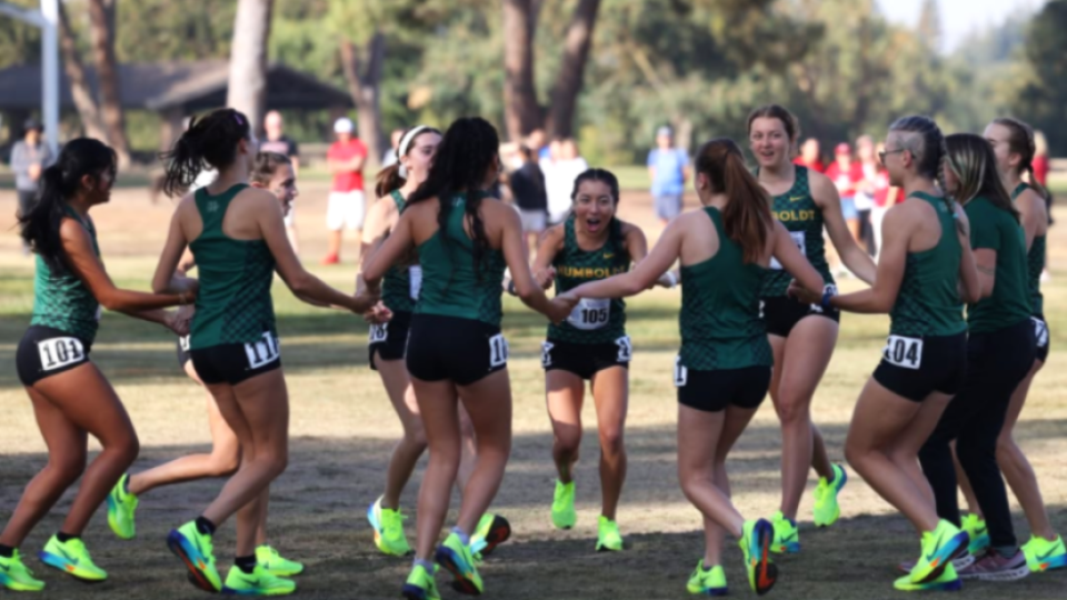 11 women's cross country teammates holding hands in a circle smiling and laughing.
