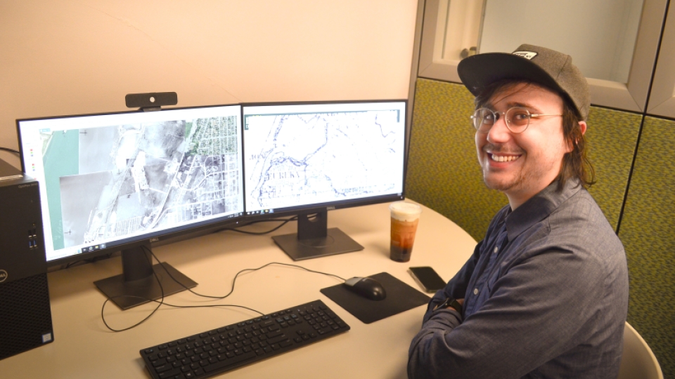 A student smiling in front of two monitors with GIS software on them.