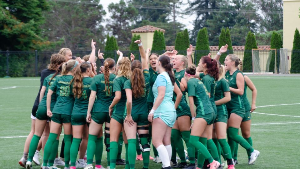 The women's soccer team in green jerseys in a huddle on the soccer field.