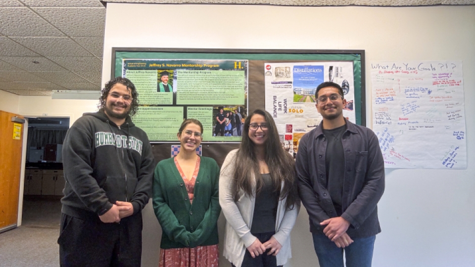 Four people smiling and standing in front of a mentorship program poster.