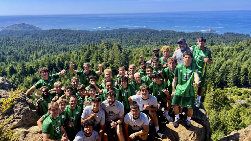 A group photo of the men's soccer team on top of a rock with the ocean in the background.