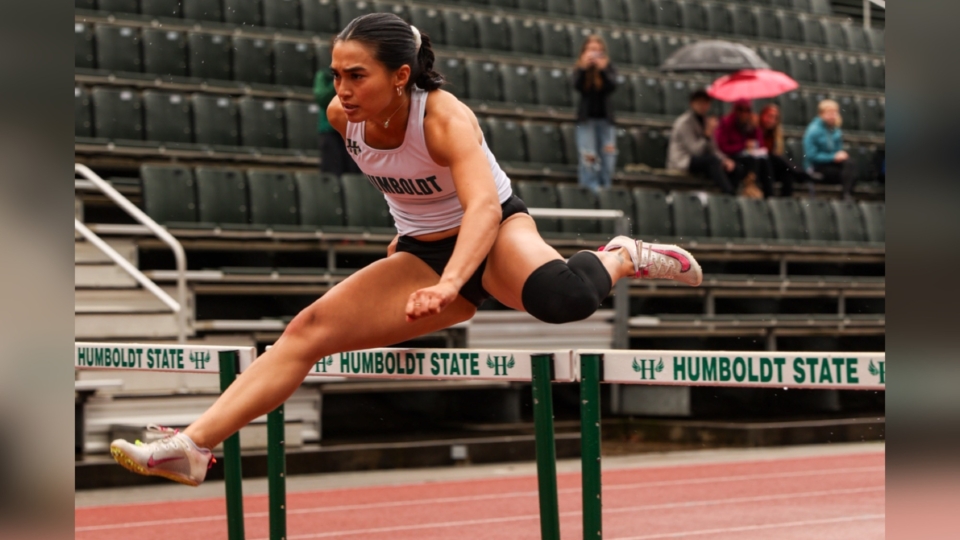 A female track athlete jumping over a hurdle.