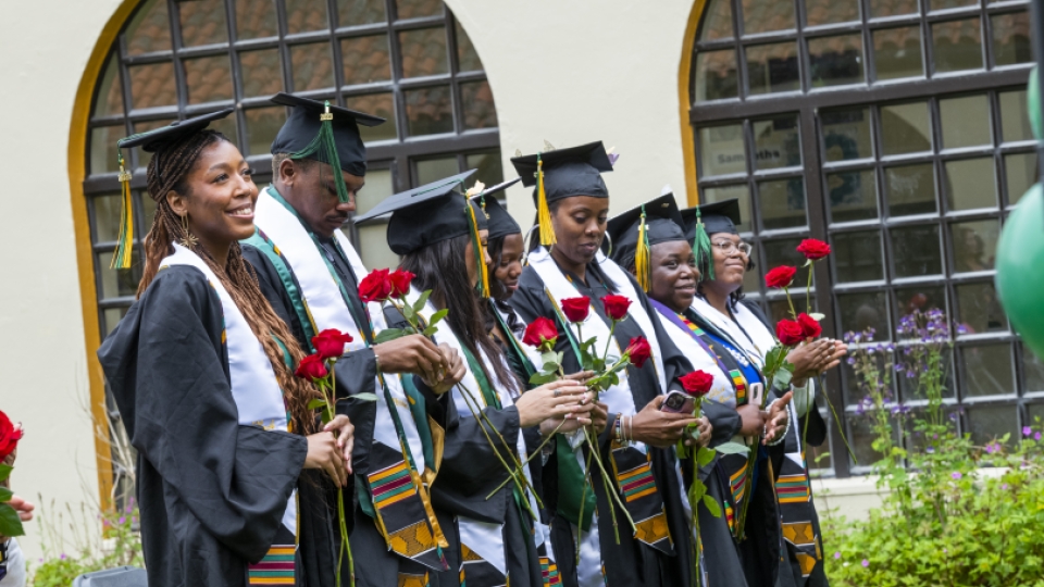 Seven Umoja grads in commencement regalia outside smiling.