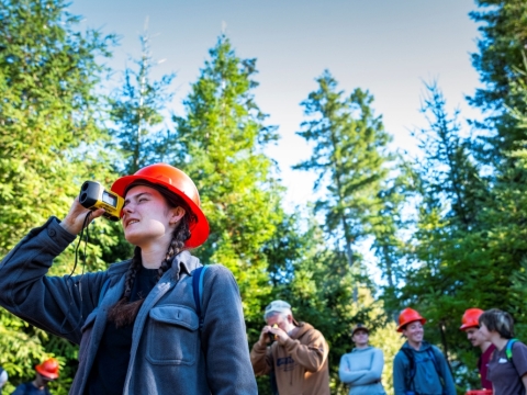 A photo of a student wearing a bright orange safety helmet, while looking through a monocular during a forestry class