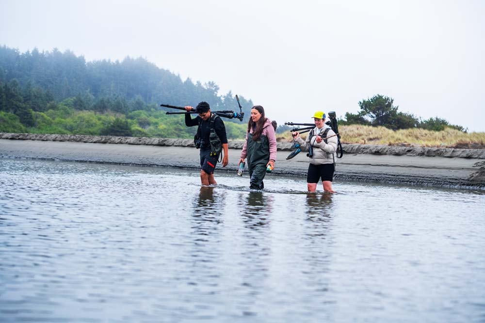 Three students wadding across body of water carrying tripods